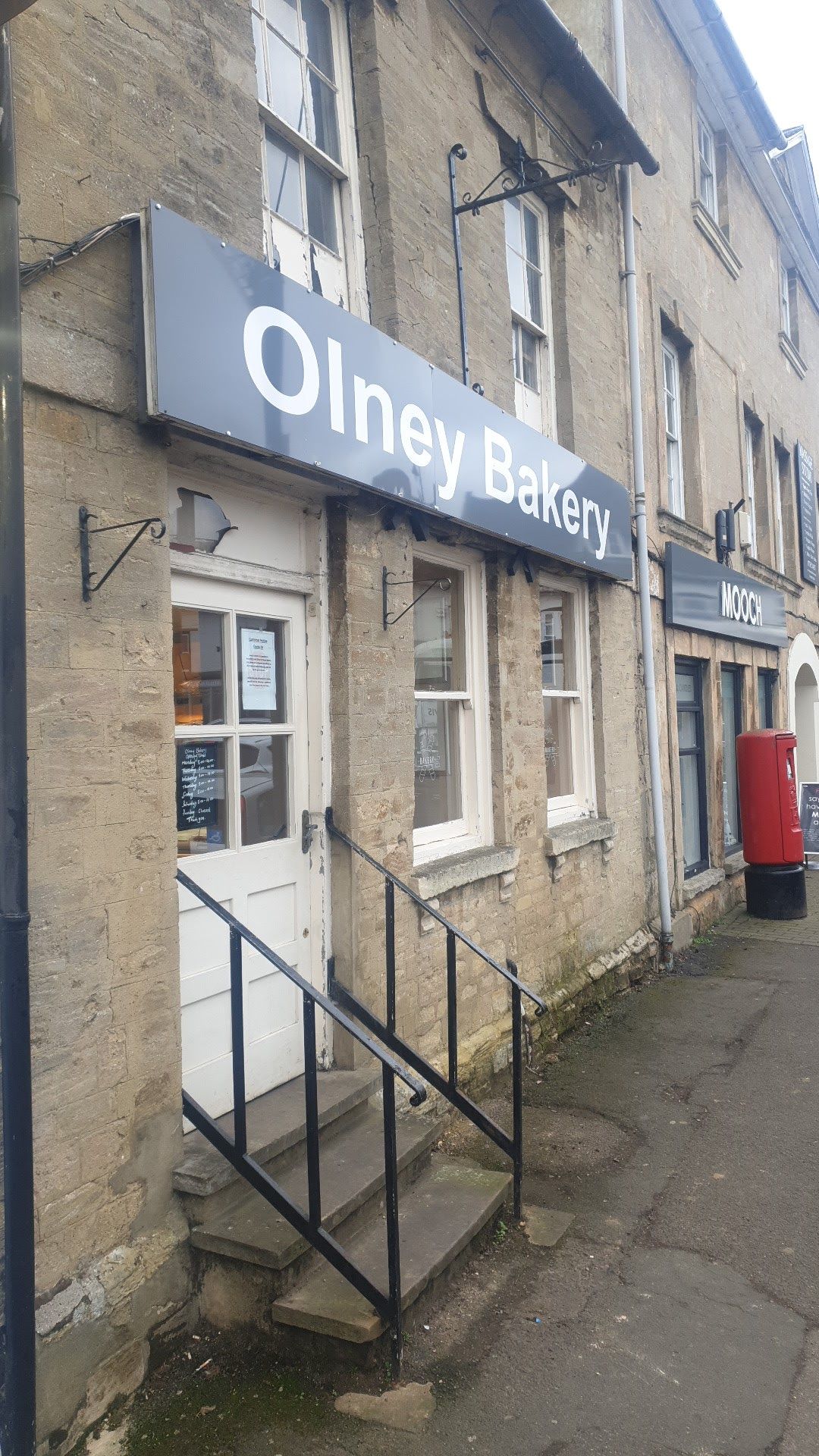 The bakery entrance: a white door at the top of three stone steps with black railings, under the Olney Bakery sign.