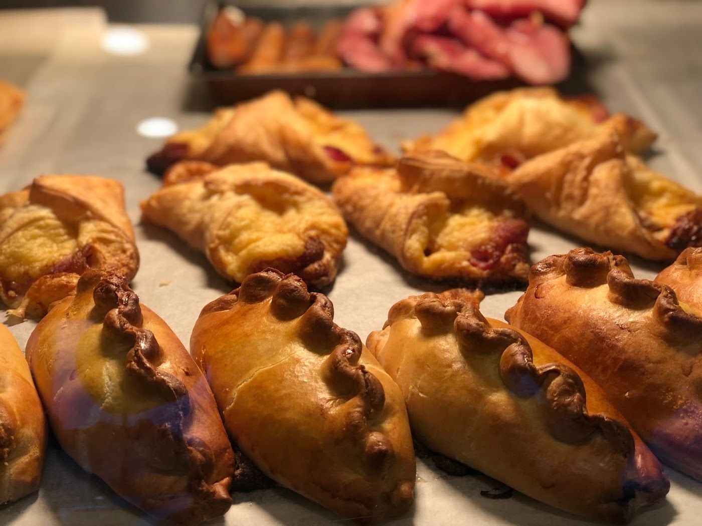 Cornish pasties with crimped edges lined up in the counter, cheese pastries behind.