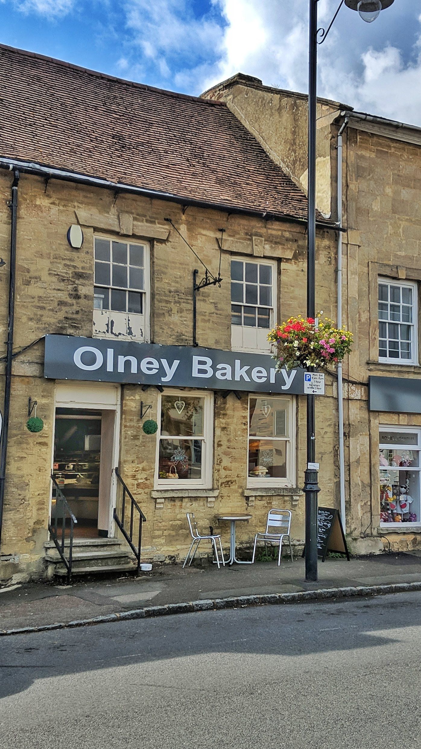 The Olney Bakery shopfront: a slate sign with white lettering on a honey limestone building, white door up stone steps with black railings, and a bistro table outside.
